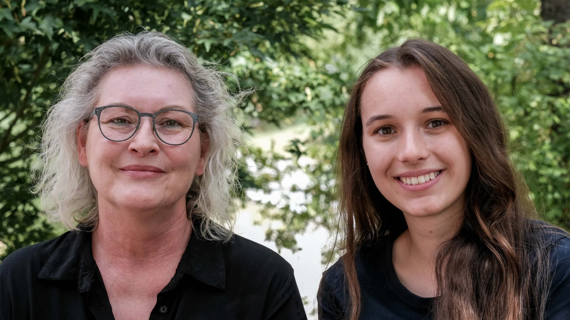 Zwei Frauen verschiedener Generationen l&auml;cheln in die Kamera vor einem Hintergrund aus gr&uuml;nen B&auml;umen und Bl&auml;ttern. Die &auml;ltere Frau tr&auml;gt eine schwarze Bluse und eine Brille, die j&uuml;ngere Frau hat lange braune Haare und ein dunkles Oberteil.