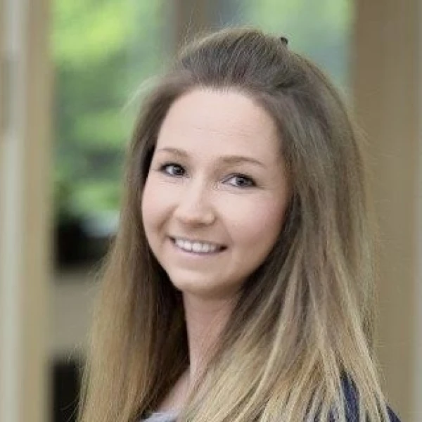 L&auml;chelnde Frau mit Schulterlangen, hellbraunen Haaren in einer grauen Bluse und blauem Blazer, die freundlich in die Kamera blickt. Der Hintergrund zeigt einen unscharfen Innenraum mit Blick nach drau&szlig;en.