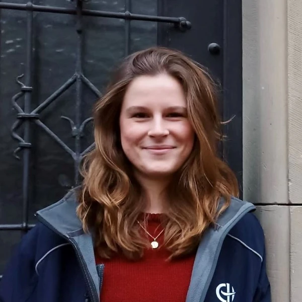 Eine l&auml;chelnde junge Frau in rotem Pullover und dunkelblauer Jacke mit Logo der Erlacher H&ouml;he steht vor einer alten dunklen Haust&uuml;re mit Glasfenster und Metallverzierung. Sie hat mittellange, gewellte braune Haare und tr&auml;gt eine Halskette.