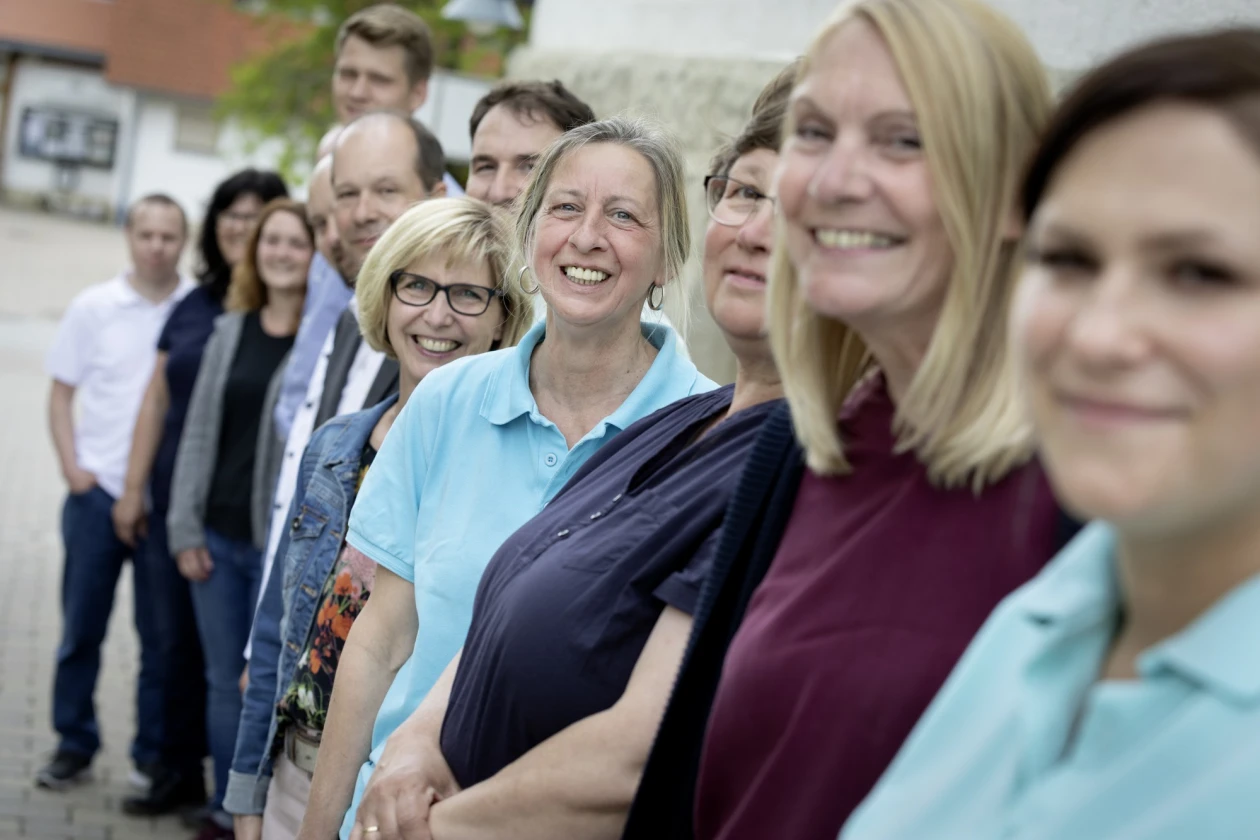 Eine Gruppe l&auml;chelnder Personen in unterschiedlicher Kleidung stehen hintereinander in einer Reihe an einer Steinmauer. Im Hintergrund ist ein wei&szlig;es Geb&auml;ude mit rotem Ziegeldach zu sehen.