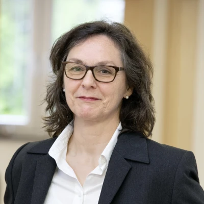 Eine l&auml;chelnde Frau mit schulterlangen braunen Haaren, mit Brille, gekleidet in einer wei&szlig;en Bluse mit schwarzem Blazer.