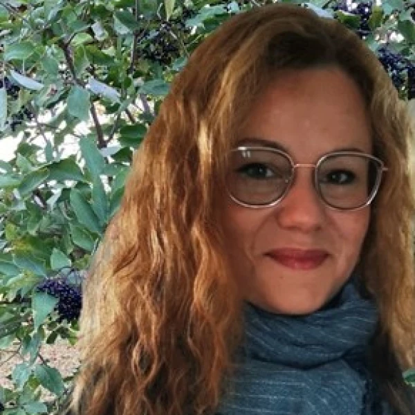 L&auml;chelnde Frau mit langem, welligen hellbraunen Haaren und Brille, mit einem blaugrauen Schal, steht vor einem Hintergrund aus gr&uuml;nen Bl&auml;ttern mit schwarzen Beeren.