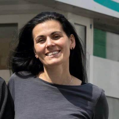 L&auml;chelnde Frau mit schwarzen Haaren in einem grauen Langarmshirt steht vor einem Geb&auml;ude.