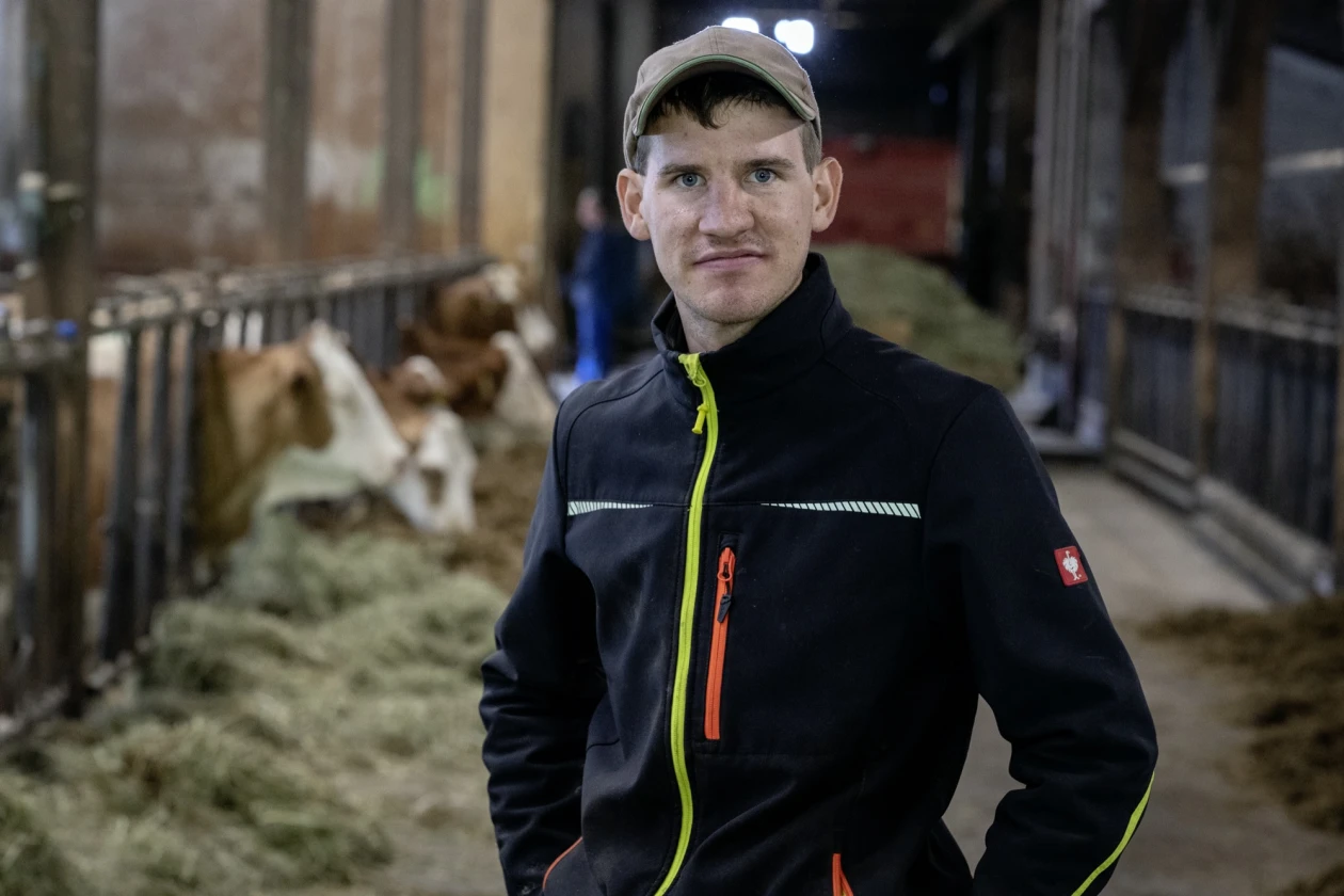 Junger Mann mit Kappe in schwarzer Arbeitsjacke mit neongelben Akzenten steht im Kuhstall. Im Hintergrund sind Rinder zu sehen. Der Mann blickt direkt in die Kamera.