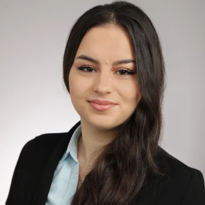 Junge Frau mit langen dunklen Haaren in einem schwarzen Blazer und hellblauer Bluse, die in die Kamera l&auml;chelt.