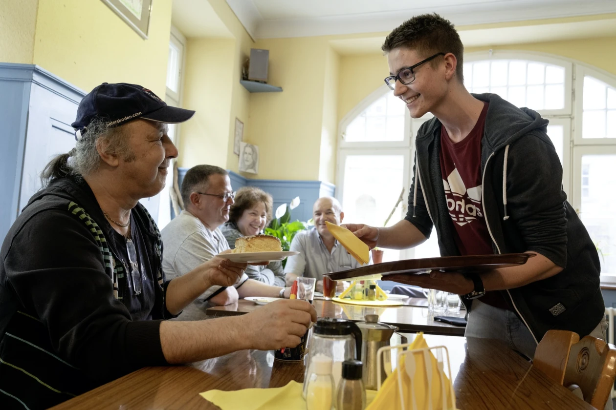 Ein junger Mann serviert einem Mann einen Kuchen an den Tisch in einem hellen Speisesaal. Mehrere Menschen sitzen im Hintergrund und kommunizieren w&auml;hrend einer Mahlzeit. Eine freundliche Atmosph&auml;re des Miteinanders und der Unterst&uuml;tzung ist sichtbar.