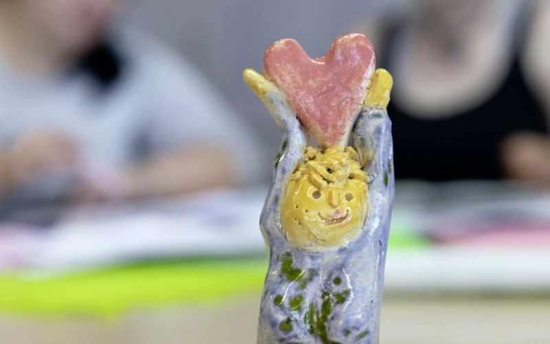 Handgefertigte Keramikfigur mit gelbem Gesicht und rosa Herz, in blau-gr&uuml;ne Glasur getaucht, die auf einem Holztisch vor verschwommenen Personen im Hintergrund steht.