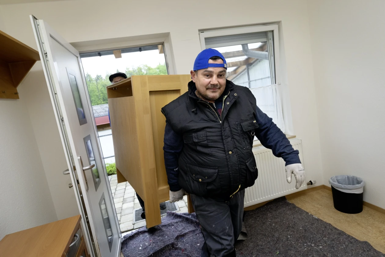 Zwei Personen tragen einen Schrank durch die ge&ouml;ffnete Eingangst&uuml;r in einen Raum. Der Mann vorne tr&auml;gt eine schwarze Weste und eine blaue Kappe, vom hinteren Mann ist nur die Kappe zu sehen.