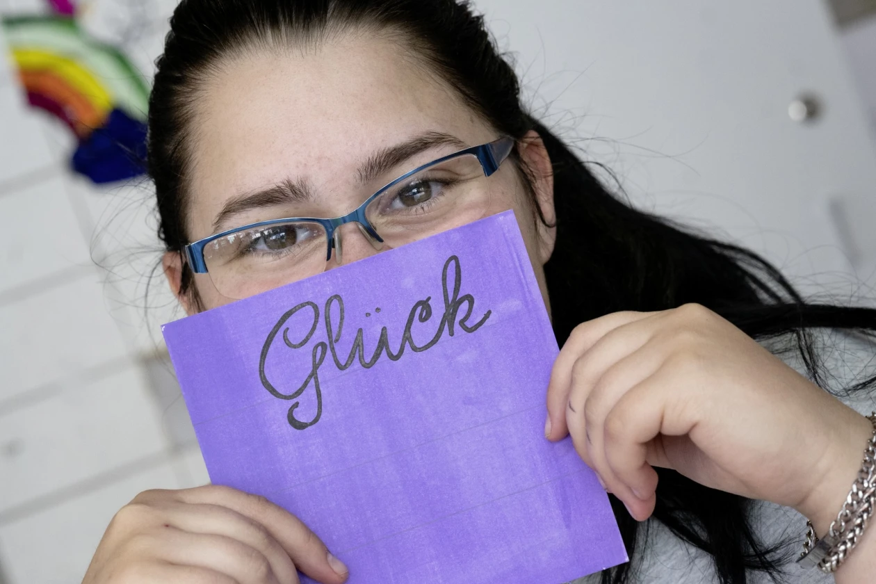 Eine junge Frau mit Brille und dunklen langen Haaren tr&auml;gt ein graues T-Shirt mit dem Aufdruck "Live the life you". Sie h&auml;lt eine lila Karte mit dem Wort "Gl&uuml;ck" vor ihr Gesicht. Sie sitzt an einem Tisch mit Dokumenten und l&auml;chelt in die Kamera.